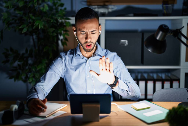 Joven hispano trabajando en la oficina por la noche haciendo un gesto de stop con las manos en las palmas, expresión de enojo y frustración  - Foto, imagen
