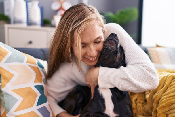Νεαρή ξανθιά γυναίκα αγκαλιάζει το σκυλί κάθεται στον καναπέ στο σπίτι - Φωτογραφία, εικόνα