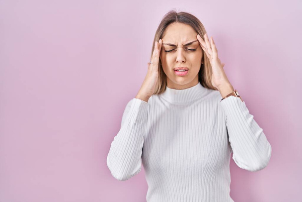 Jeune femme blonde portant un pull blanc sur fond rose avec la main sur la tête pour la douleur à la tête parce que le stress. migraine souffrant.  - Photo, image