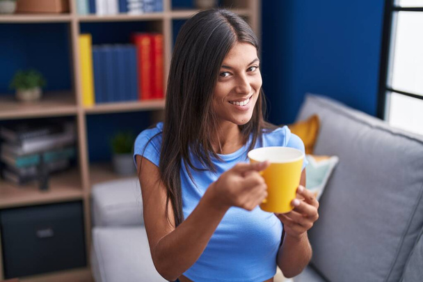 Nuori kaunis latino nainen juo kahvia istuu sohvalla kotona - Valokuva, kuva