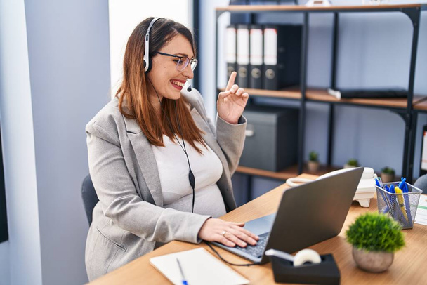 Pregnant woman working at the office wearing operator headset smiling happy pointing with hand and finger to the side  - Фото, изображение