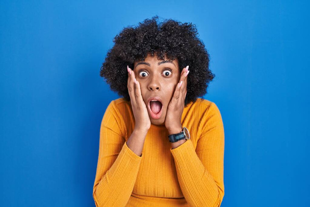 Zwarte vrouw met krullend haar staan over blauwe achtergrond bang en geschokt, verrassing en verbaasd uitdrukking met handen op het gezicht  - Foto, afbeelding