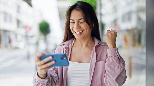 Young beautiful hispanic woman smiling confident watching video on smartphone celebrating at street - Photo, image