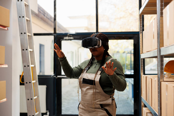 Storage room employee wearing virtual reality headset, enjoying warehouse simulation, preparing customers orders in storehouse. African american supervisor working at merchandise inventory - Photo, Image