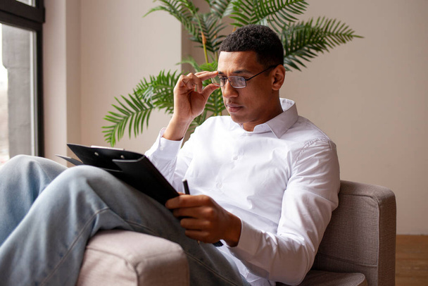 зайнятий серйозний афроамериканський чоловік у сорочці і окулярах сидить на стільці і пише в буфері, хлопець пише ручкою на папері і налаштовує окуляри - Фото, зображення