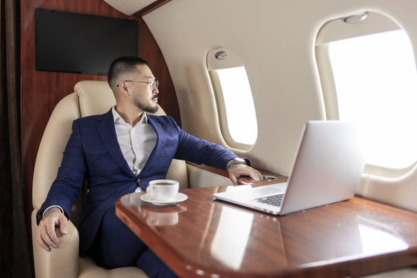 successful Asian businessman in suit and glasses flies in private luxury plane with laptop, Korean trader financier works at computer in airplane, luxury lifestyle - Photo, Image