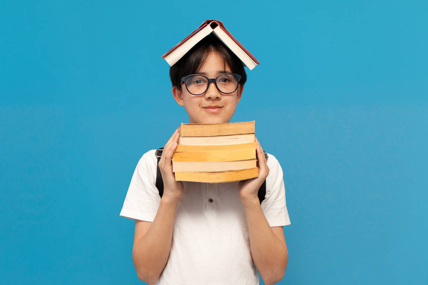 asian boy schoolboy of twelve years old wearing glasses and with backpack holds lot of books on blue isolated background, korean child goes back to school to study and learns lessons - Fotó, kép