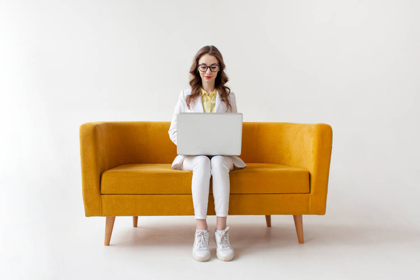 Takım elbiseli genç iş kadını rahat bir kanepeye oturur ve dizüstü bilgisayar kullanır. Resmi giysili kız bilgisayardaki sarı kanepede beyaz izole edilmiş arka planda daktilo yazar. - Fotoğraf, Görsel