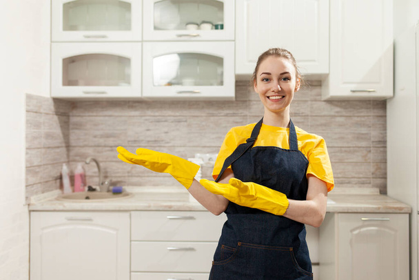vrolijke huishoudster in schort en handschoenen reinigt huizen en glimlachen, vrouw huisvrouw in uniform staat tegen de achtergrond van de keuken en adverteert kopieerruimte - Foto, afbeelding