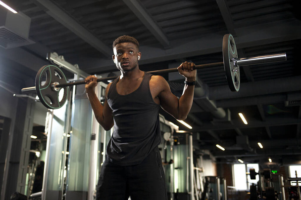 joven afroamericano hombre levanta barbell en oscuro gimnasio, chico deportivo entrena en sala de fitness y entra para deportes - Foto, imagen