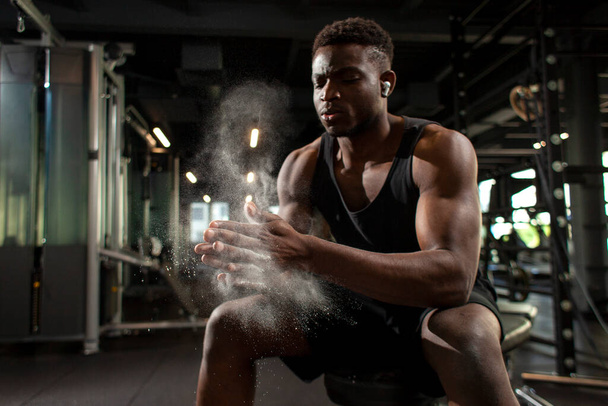 sporty african american man trains in dark gym and rubs his hands with magnesia, young guy makes clap with his hands in the fitness room, close-up of dust and flour with the hands of athlete - Фото, изображение