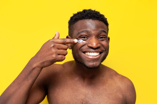 african american man applies skin cream on his face and smiles on yellow isolated background, the guy takes care of his face skin and uses cosmetics - Fotó, kép