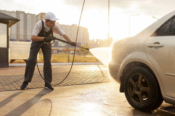 jonge man in uniform auto wassen werknemer wast auto met water uit slang, man in overall werkt in openlucht auto schoonmaak service - Foto, afbeelding