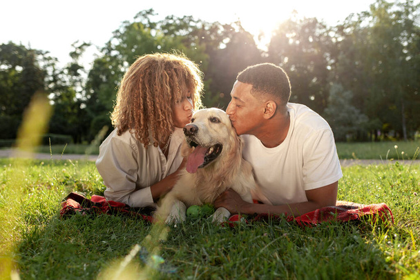 young african american couple with dog lie in park on green grass, curly woman and young man kiss and love golden retriever, young happy family with pet in nature - Foto, Imagen