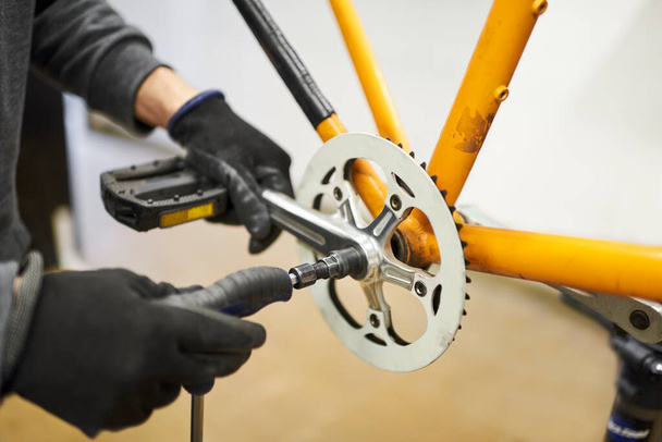 Уход за велосипедом: руки неузнаваемого человека в перчатках разбирают педали оранжевого велосипеда в мастерской. - Фото, изображение