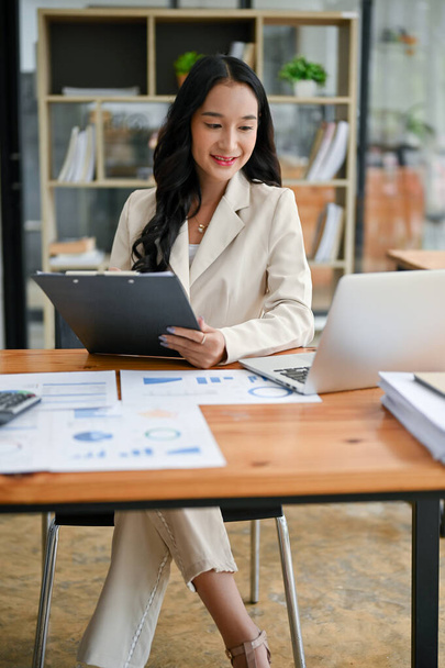 A portrait of a beautiful millennial Asian businesswoman or female executive manager in a trendy suit focuses on business reports and working on her tasks on her laptop at her desk in the office. - Foto, Imagen