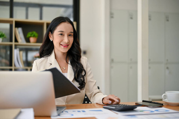 Eine attraktive asiatische Geschäftsfrau oder Buchhalterin sitzt mit Geschäftsberichten und Laptop an ihrem Schreibtisch im Büro. - Foto, Bild