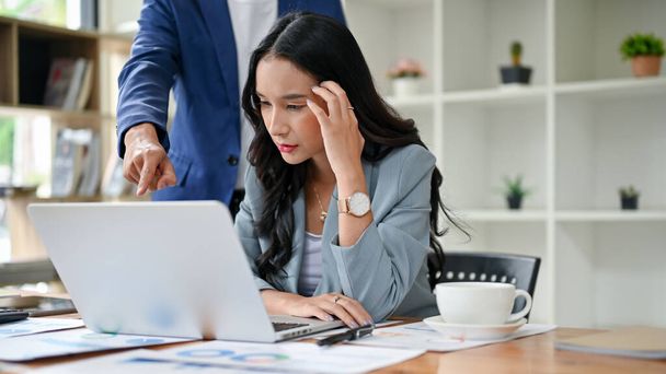 A stressed millennial Asian female office worker is being complained about and pointed finger at her laptop screen by her male boss at her desk in the office. project failure, upset, problems - Foto, Imagem