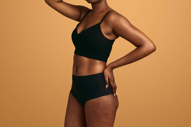 Crop mujer africana anónima en ropa interior negra con la mano en la cintura de pie contra fondo naranja concepto de amor propio y aceptación - Foto, imagen