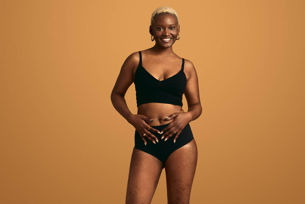 Cheerful young African American female model in black lingerie with short hair looking at camera and smiling, against beige background while touching belly in studio - Foto, Bild