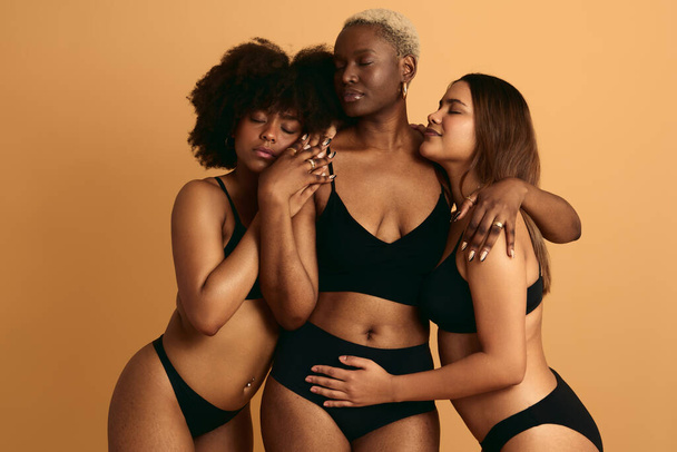 Feminine young multiethnic female plus size models in black underwear embracing with closed eyes against beige background - Fotografie, Obrázek