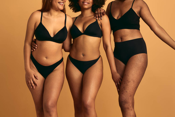 Faceless young multiracial female models in black underwear standing close to each other and hugging while smiling against yellow background in studio - Φωτογραφία, εικόνα