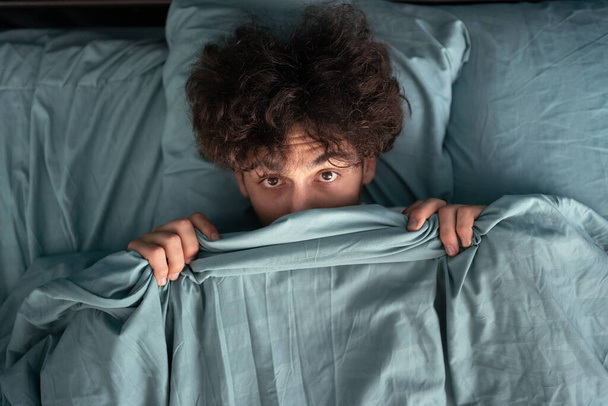 Handsome surprised guy covering half of face with blanket, scared man peeking from duvet, afraid of night monsters, top view. Close-up - Fotó, kép
