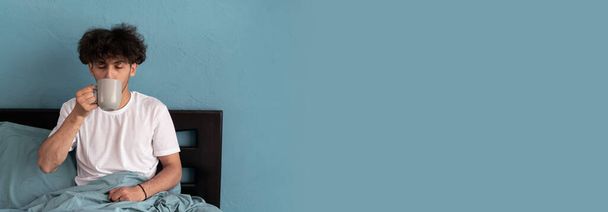 Hombre joven y guapo árabe disfruta del café por la mañana mientras está sentado en la cama vestido con pijama. Buenos días y buen concepto de humor. Banner, espacio de copia - Foto, imagen