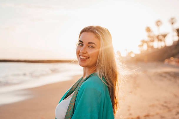 Πορτρέτο μιας χαρούμενης όμορφης γυναίκας στην άμμο της παραλίας απολαμβάνοντας και διασκεδάζοντας στο ηλιοβασίλεμα της ημέρας. Κοιτάζοντας την κάμερα χαμογελώντας. - Φωτογραφία, εικόνα