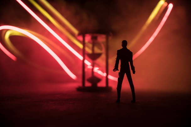 Concepto de tiempo. Silueta de un hombre parado entre clepsidras con humo y luces sobre un fondo oscuro. Imagen surrealista decorada - Foto, Imagen