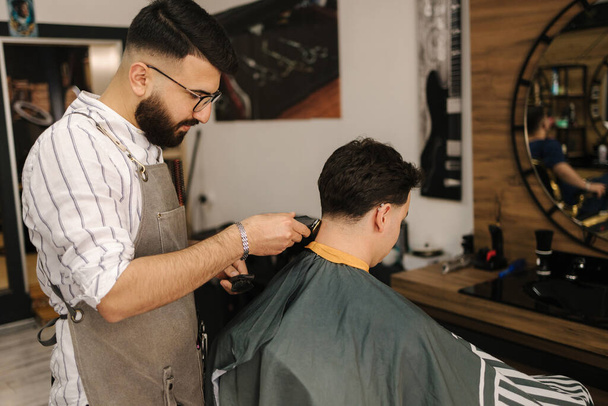 Bearded barber cutting hair of male customer. Hairstylist serving client at barber shop. High quality photo - Photo, Image