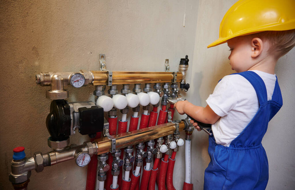 Μωρό αγόρι σε κράνος κατασκευή ασφαλείας χρησιμοποιώντας εργαλείο κλειδί, ενώ για τον καθορισμό υδραυλικών εγκαταστάσεων με σωλήνες και μετρητή θερμόμετρο. Παιδί υδραυλικός επισκευή υδραυλικό σύστημα σωλήνων στο διαμέρισμα υπό ανακαίνιση. - Φωτογραφία, εικόνα