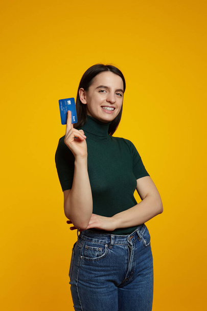 Vertical photo of cheerful caucasian girl wearing green shirt, hold in hand mock up of credit bank card isolated on plain yellow background - Foto, afbeelding