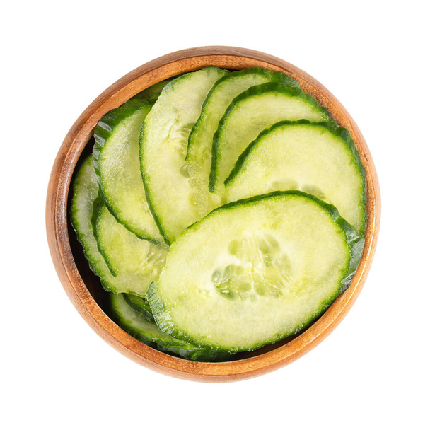 Cucumber slices, in a wooden bowl. Fresh salad cucumber, with green skin, diagonally sliced unripe fruit of Cucumis sativus, a vegetable. Close-up, from above, isolated, over white, macro food photo. - Foto, immagini