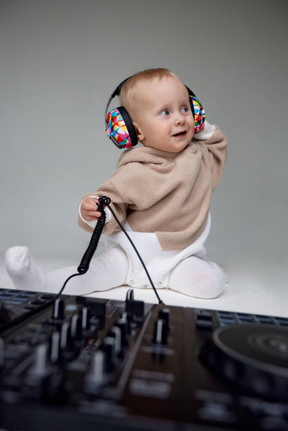 Ein niedliches, lächelndes blondes Baby in einem beigen Kapuzenpulli sitzt auf dem Boden und spielt mit DJ-Kopfhörern und einem DJ-Mischpult. Musik und Spaß. Isoliert auf grauem Hintergrund - Foto, Bild