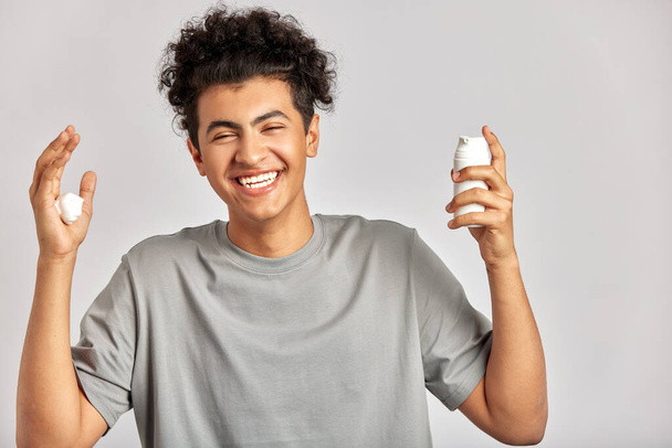 Young handsome smiling guy with black curly hair squeezing skincare cream from the dispenser to his hand. Biracial man in studio with healthy skin procedure. Wellness, cosmetics and skincare for men - Foto, Imagem
