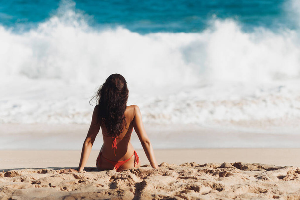 Bikini beach woman sitting by the ocean on a sandy beach. Rear view of brunette Caucasian girl model looking at the waves. The concept of a romantic tropical vacation - Φωτογραφία, εικόνα