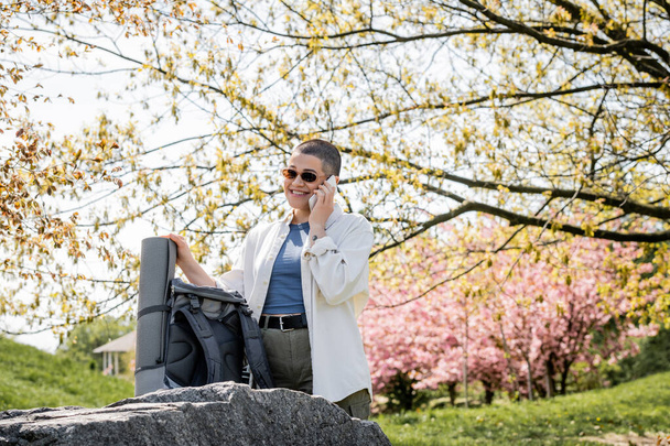 Usmívající se krátkovlasá a tetovaná turistka ve slunečních brýlích mluvící na smartphonu a stojící u batohu na kameni s malebnou přírodou na pozadí, sebevědomá průzkumnice - Fotografie, Obrázek