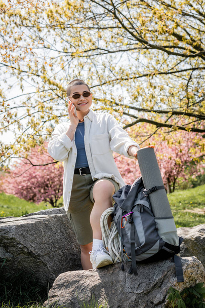 Positive junge kurzhaarige Touristin in lässiger Kleidung und Sonnenbrille, die mit dem Smartphone spricht, während sie in der Nähe von Rucksack und Steinen mit der Natur im Hintergrund steht, selbstbewusste Forscherin - Foto, Bild