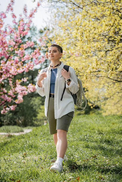 Smiling short haired and tattooed female hiker in casual clothes holding backpack while walking on grass with landscape at background, independent traveler embarking on solo journey - Φωτογραφία, εικόνα