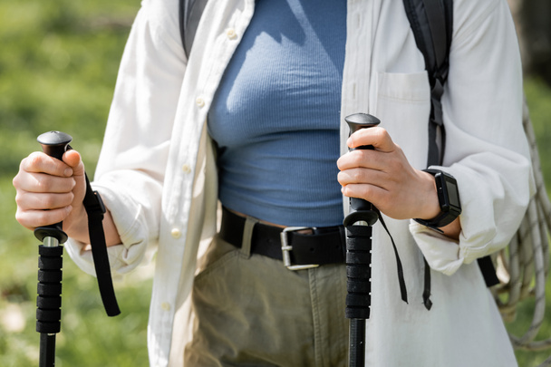 Oříznutý pohled na mladou ženu turistka v ležérním oblečení s batohem drží trekking tyče při chůzi venku, nezávislý cestovatel nastupující na sólo cestu - Fotografie, Obrázek
