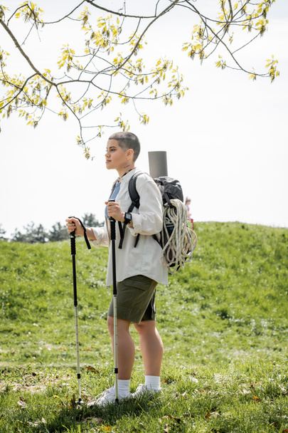 Jóvenes viajeras de pelo corto con ropa casual con mochila y equipo de viaje caminando con bastones de trekking con paisaje de fondo, viajeras independientes embarcándose en un viaje en solitario - Foto, Imagen