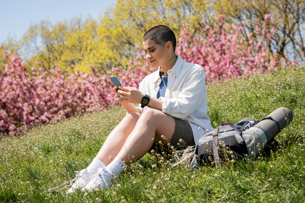 Lächelnde junge kurzhaarige Reisende mit Smartphone, während sie in der Nähe eines Rucksacks mit Reiseausrüstung auf einem grasbewachsenen Hügel mit der Natur im Hintergrund sitzt, neugierige Wanderer, die neue Landschaften erkunden - Foto, Bild