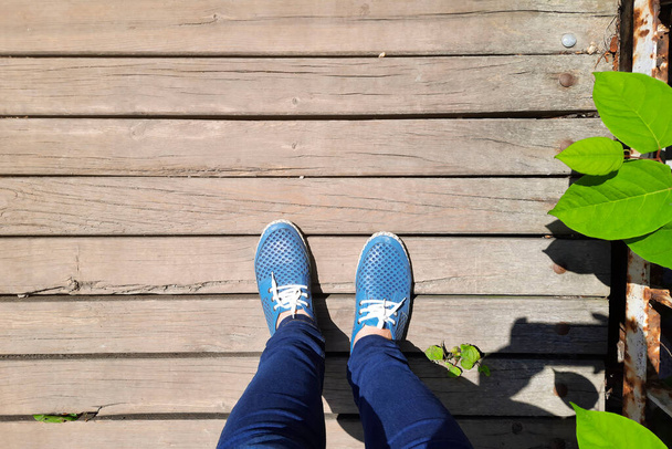 View from above on legs in moccasins on background of old wooden bridge. Women's feet in blue shoes and jeans stand on wooden plank road. Country walk on sunny day. - Photo, Image