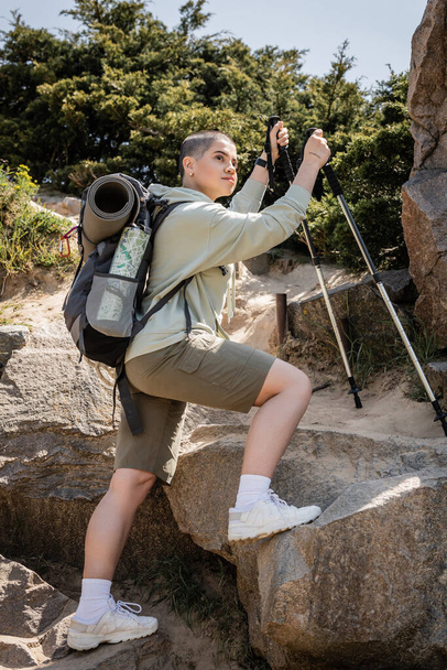 short haired female traveler with backpack and map holding trekking poles while standing on stones near hill at background, tranquil hiker finding inner peace on trail, Translation of tattoo: love - Photo, Image