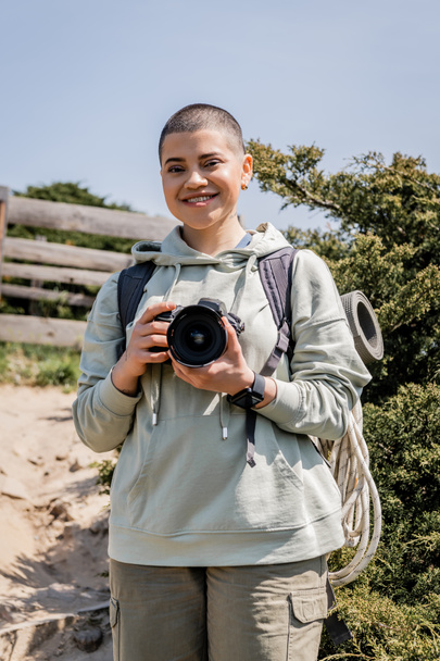 Cheerful young short haired female tourist with backpack and travel supplies holding digital camera and looking at camera with nature at background, hiker finding inspiration in nature - Photo, Image