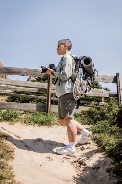 Oldalnézet fiatal rövid hajú női túrázó hátizsákkal és utazási felszereléssel digitális fényképezőgép és álló dombon ég a háttérben, túrázó inspirációt a természetben - Fotó, kép