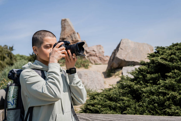若い短い髪とバックパックと地図と入れ墨の女性観光客は、バックグラウンドで自然と一緒に立っている間、デジタルカメラで写真を撮る,タトゥーの翻訳:愛 - 写真・画像