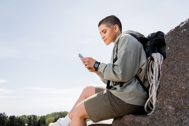 Młoda, krótkowłosa kobieta podróżująca z plecakiem i sprzętem podróżującym za pomocą smartfona, siedząca na kamieniu z naturą i błękitnym niebem w tle, żywe doświadczenia z podróży, lato - Zdjęcie, obraz