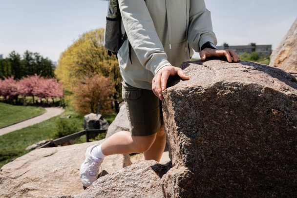 Cropped view of young female hiker with backpack and fitness tracker standing near stones on hill with blurred nature at background, vibrant travel experiences, summer - Photo, Image